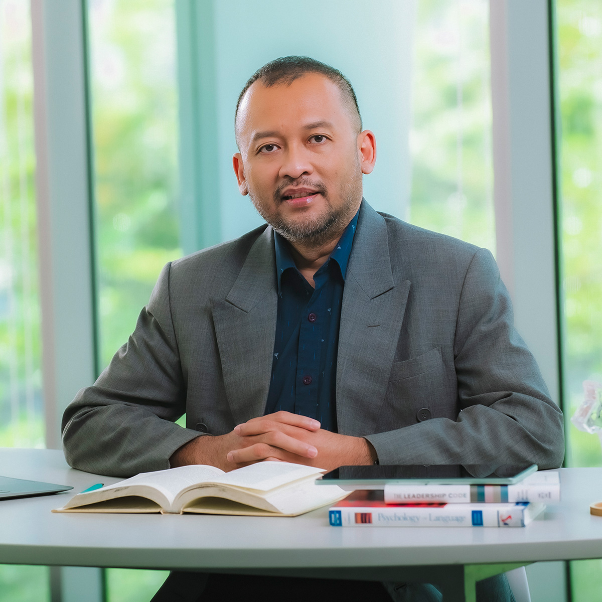Dr Teguh Haryo Sasongko seated at a desk with books, wearing a grey jacket